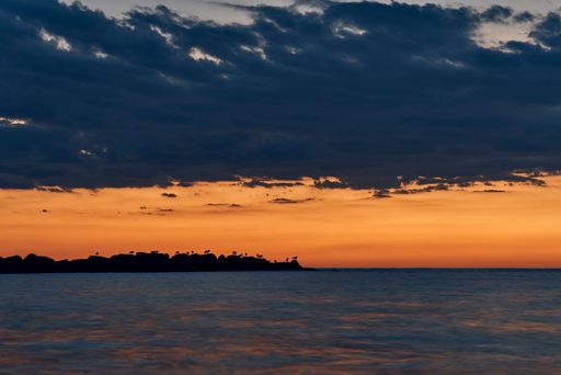 Serene sunset over the ocean with silhouetted rocks and birds against a vibrant orange sky.