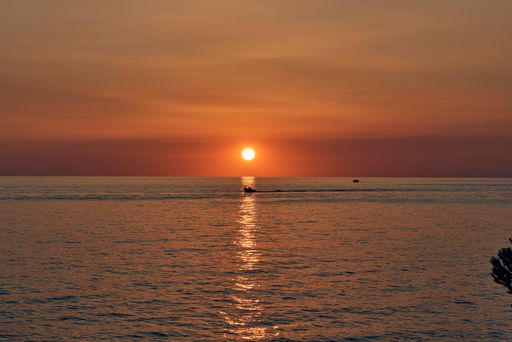 Sunset over a calm sea with a boat silhouette and vibrant orange sky reflecting on the water.