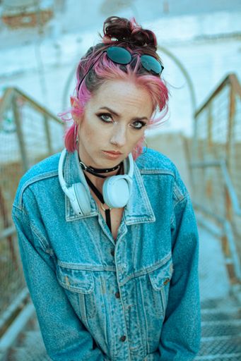 A vertical shot of a cool white lady with pink hair, headphones, gothic style makeup and clothing