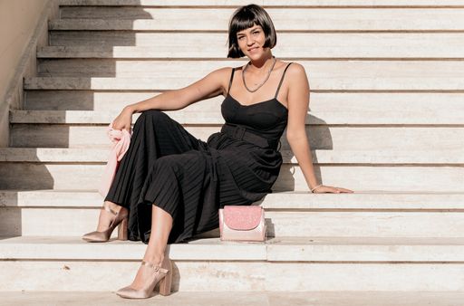 Front view of stylish young woman wearing elegant black jumpsuit and pink handbag, sitting on stairs, looking at camera