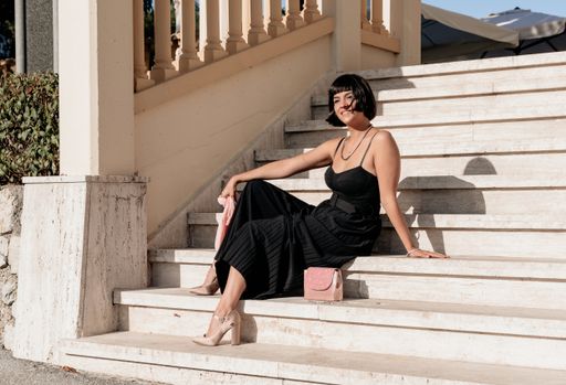 Front view of stylish young woman wearing elegant black jumpsuit and pink handbag, sitting on stairs, looking at camera