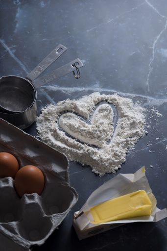Ingredients for baking with a heart drawn in flour on a dark marble surface, including eggs, butter, and measuring cups.