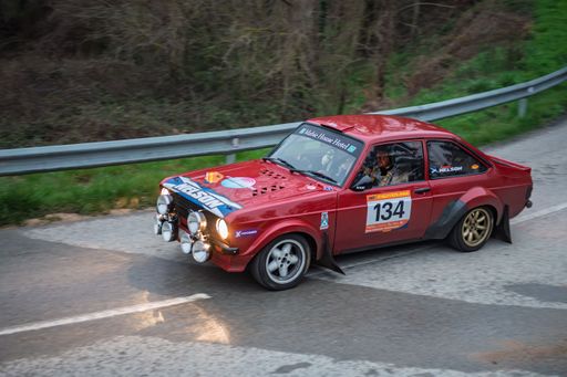 Craig Nelson and Willie Nelson driving a red Ford Escort RS1600 with headlights on during the 73 Rally Costa Brava, captured from a side view at dusk.