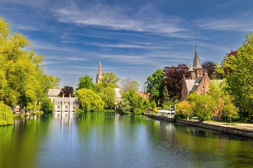 Scenic view of Bruges, Belgium with historic buildings and lush greenery reflecting in a tranquil lake under a vibrant blue sky.