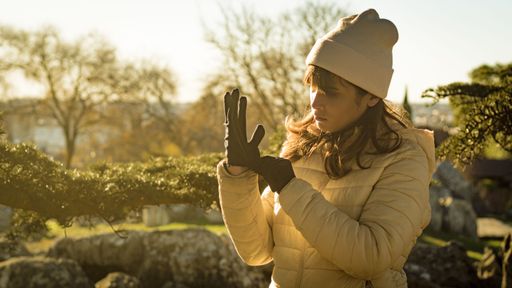 A woman in her 30s wearing a beanie and gloves enjoys a sunny autumn day in a park, surrounded by trees and rocks.