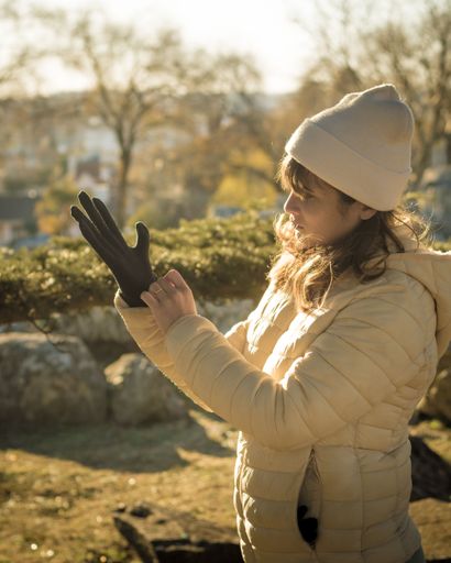 Young woman in winter coat and beanie putting on gloves in a sunny park setting.