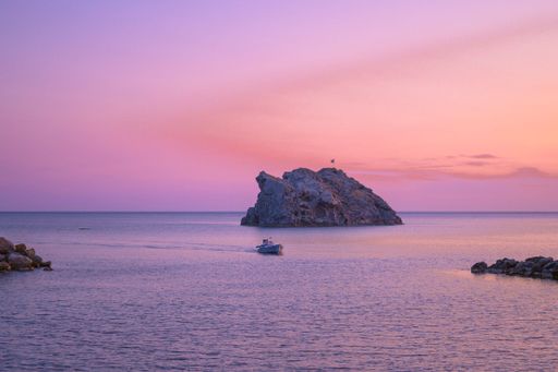 A serene seascape at sunset with a boat near a rocky island under a vibrant pink and purple sky.