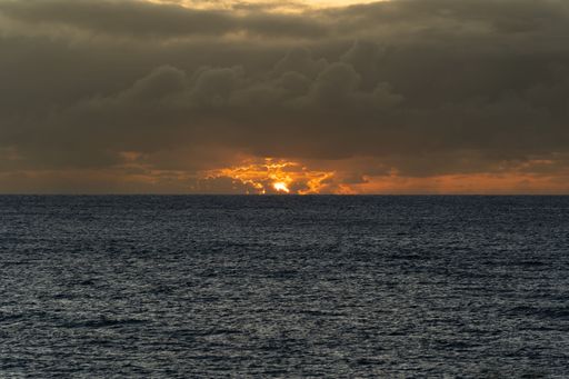 A dramatic ocean sunset with dark clouds and vibrant orange sky reflecting on the water horizon.