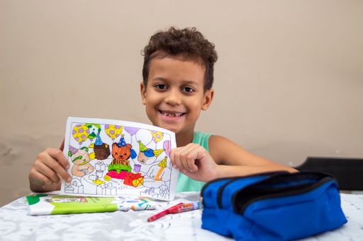 Smiling young boy holding a colorful drawing at a table with art supplies, showcasing creativity and happiness.