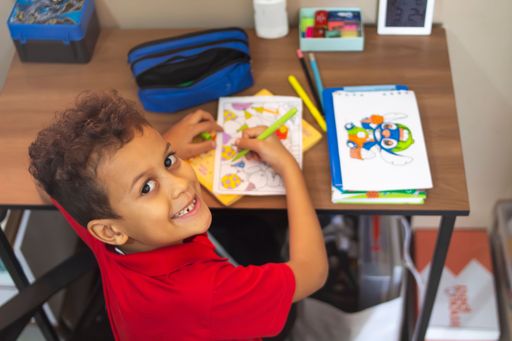 A young boy in a red shirt coloring at a desk with art supplies, smiling at the camera.