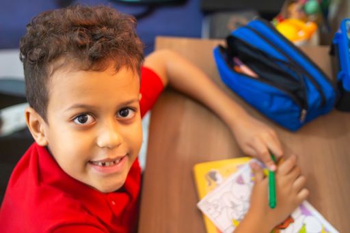 A young boy in a red shirt coloring at a desk with a blue pencil case, smiling at the camera.