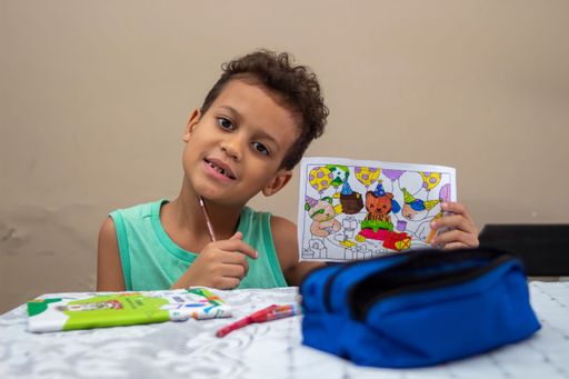 A young boy, around 6-8, proudly displays his colorful drawing while sitting at a table, showcasing creativity and joy in a casual setting.
