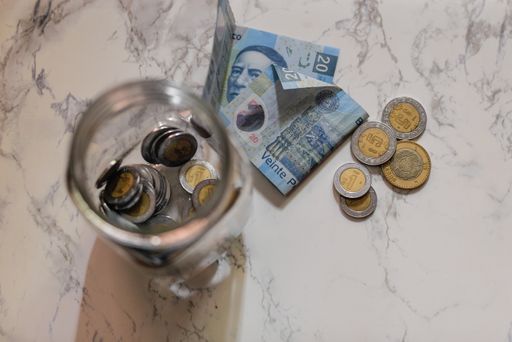 A high angle shot of a jar with coins on the table near pesos under the lights - money concept