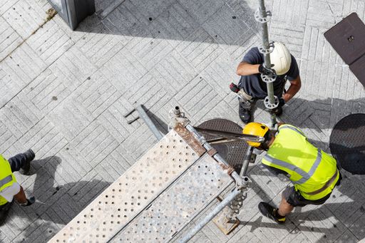 Two construction workers assembling european scaffolding on a city street sidewalk.