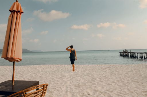 A lone person walking on the sandy beach with an umbrella and wicker chair on the foreground and the ocean in the background