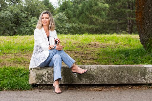 Middle-aged woman sitting on a stone bench in a park.The woman is wearing jeans, a white jacket and shows glasses in her hands.
