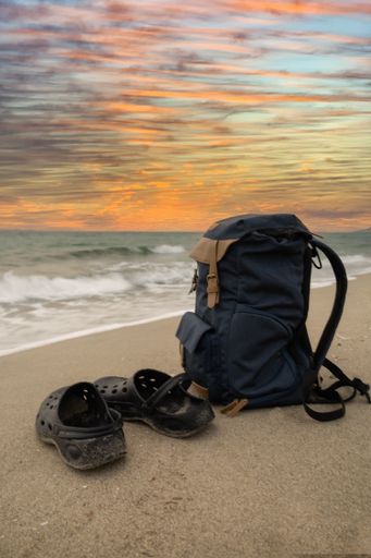 A vertical closeup shot of rubber slippers and a backpack on the seashore with waves in the background