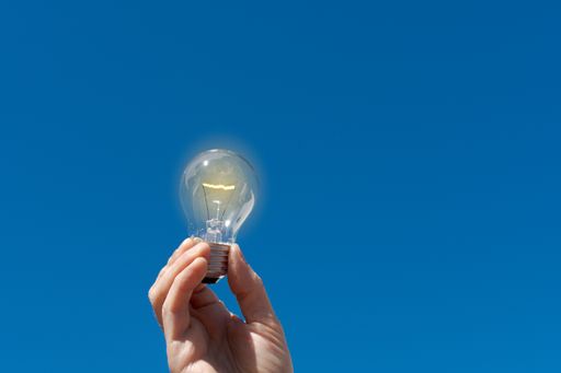 A hand holding an illuminated electric light bulb with the blue sky in the background