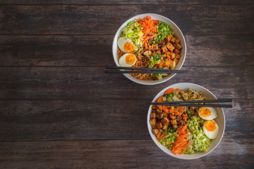 Variation of ramen homemade soup with noodles, marinated tofu, chopped carrots, onions, lettuce and broccoli in in two bowls on a wooden table.