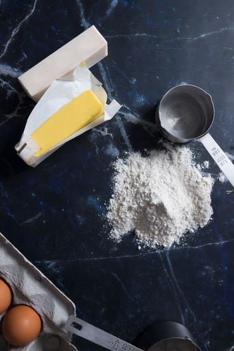 A top view of baking ingredients including butter, flour, and eggs on a dark marble surface.