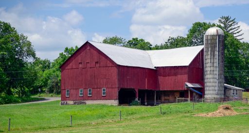 A rustic red barn with a silo in a lush green field under a blue sky, capturing rural life.