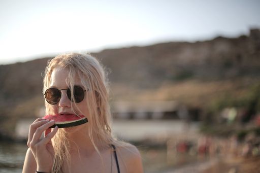 A beautiful young blonde female with sunglasses eating watermelon on the beach