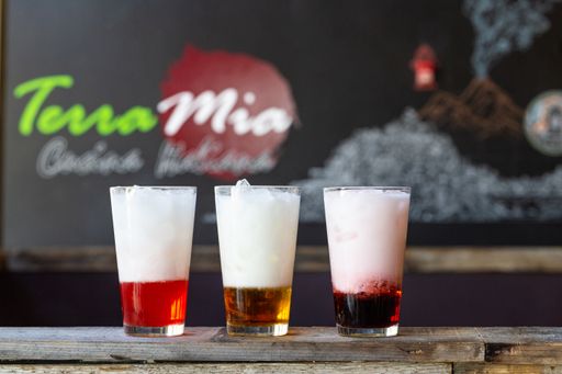 Shallow focus shot of red, yellow and purple beverages with ice and foam on wooden counter, blurry background with Terra Mia and volcano image on wall