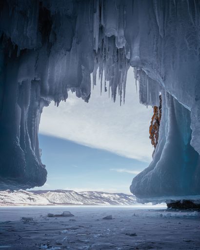 A breathtaking view through icicles of a frozen lake and snowy mountains under a clear blue sky.
