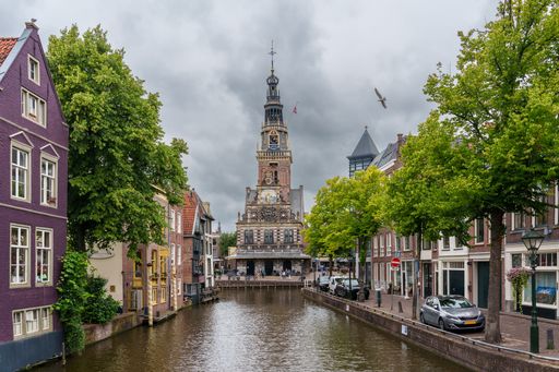 Scenic view of Alkmaar's historic Cheese Market building along a canal with lush trees and overcast sky.