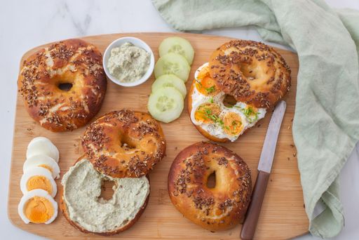 A top view of freshly baked whole grain bagels, sliced eggs, and cucumbers on a wooden board