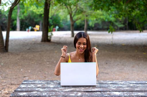 Young woman in her 20s using a laptop outdoors in a park, expressing excitement and joy, surrounded by lush green trees.