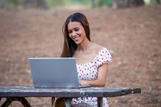 Young woman in a floral dress using a laptop outdoors, sitting on a wooden bench in a park, smiling and engaged.