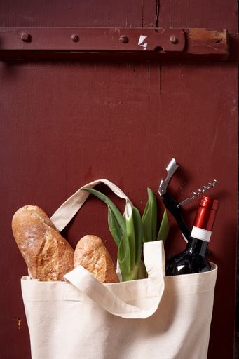 A vertical shot of wine, baguettes, corkscrew, and green beans in a tote bag on a red background