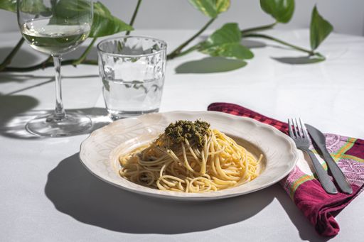 A serving of spaghetti with pesto sauce, a glass of white wine, water, and cutlery on a white table