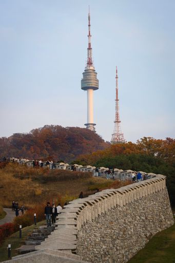 A Namsan Seoul Tower rises above autumn foliage and a historic stone wall with people walking, capturing a blend of nature and architecture.