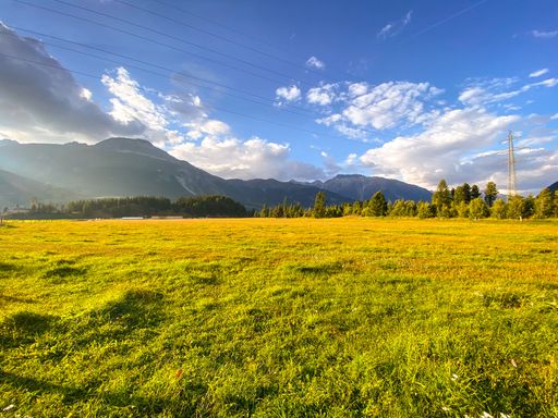 A beautiful green field surrounded by mountains and trees under a blue cloudy sky on a sunny day