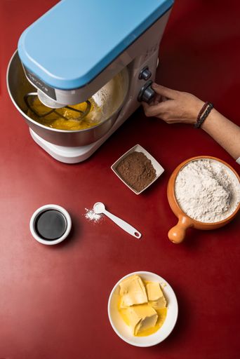 A vertical shot of an electric mixer, flour, butter, cocoa, and other ingredients on a red table