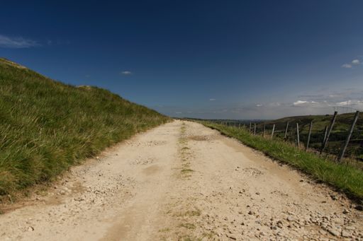 A narrow road surrounded by hills covered in greenery under the sunlight and a blue sky