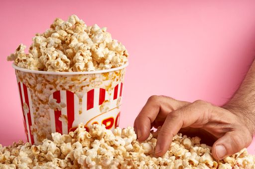 A man's hand holding popcorn and bucket of candy popcorn on pink background