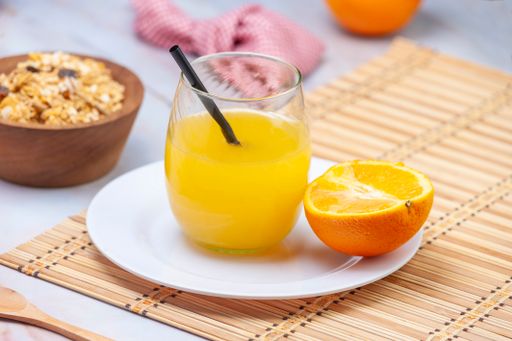 A closeup of fresh orange juice in the glass with a straw and a piece of orange on a white plate with cereals on the table