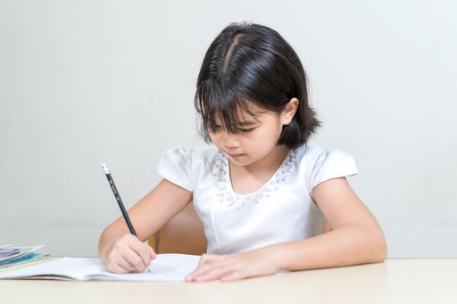 An asian little girl sitting by a desk and writing in her notebook against a white background