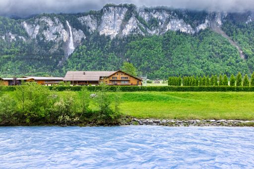 Scenic view of a riverside house with lush greenery and mountains in the background on a cloudy day.
