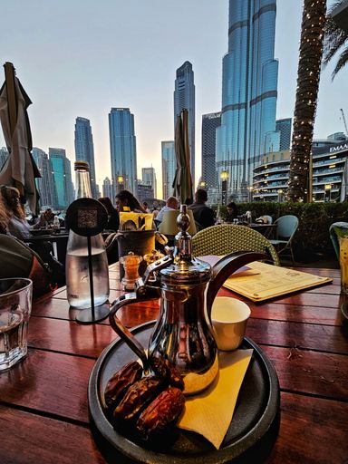 Outdoor evening scene shows a traditional metal Arabic coffee pot, dates, and a cup on a table, with the Dubai skyline visible in the background.