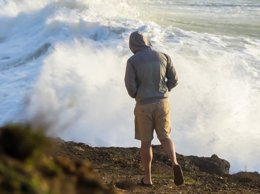 A man wearing a gray jacket with a hood from behind looking at the ocean from the cliff
