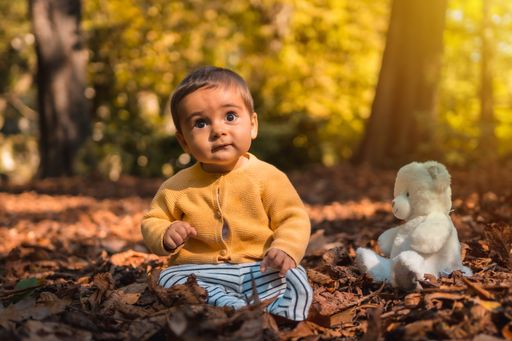 A cute six-month-old baby with a white teddy bear sitting among fallen autumn leaves in a park