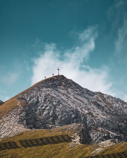 A vertical shot of two people climbing near the cross on the top of a summit