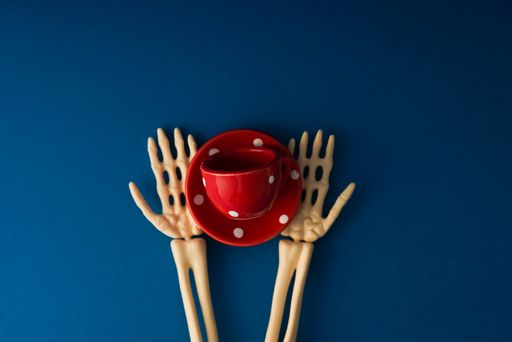 A top view of skeleton hands holding a red cup on a saucer against a blue background