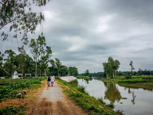 Two people walking along a pathway on the lake coast surrounded by trees under a cloudy sky