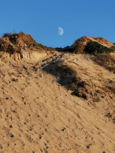 A vertical shot of bare mountains in the dune before sunset with a visible moon