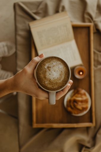 A man holding a cup of coffee on a blurred book and croissant on a wooden tray